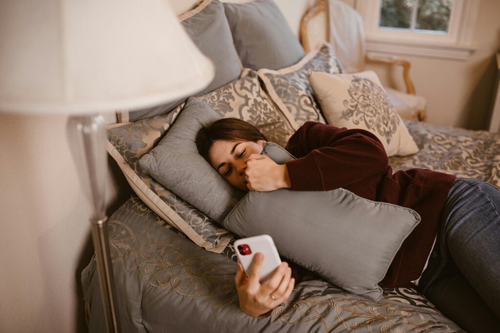 A young woman lying on a bed, holding a phone and a pillow, symbolizing relaxation and connection.